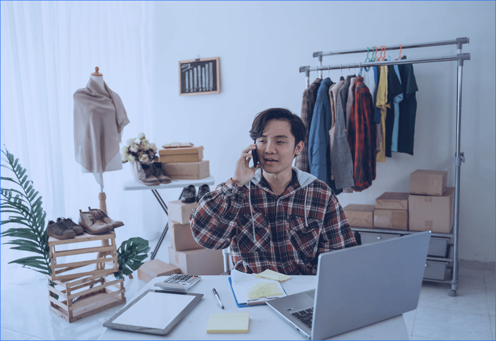 Small business owner managing an online clothing store, talking on phone while working on a laptop with inventory and packaging boxes around.